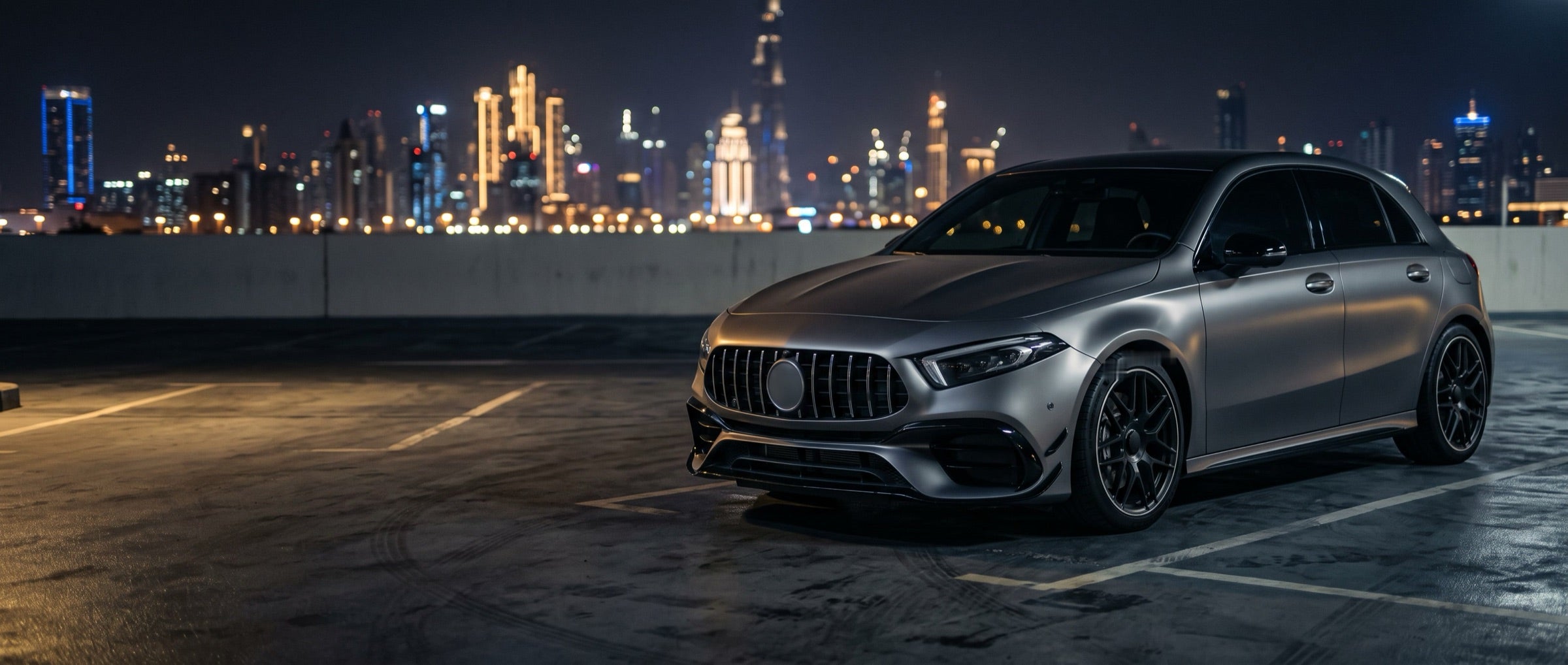 Grey hatchback on a rooftop car park with a city skyline at night